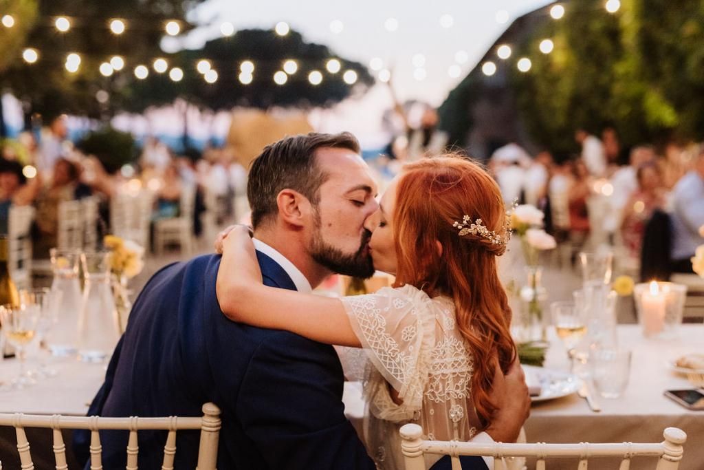Una sposa e uno sposo si baciano mentre sono seduti a un tavolo durante un ricevimento di nozze.