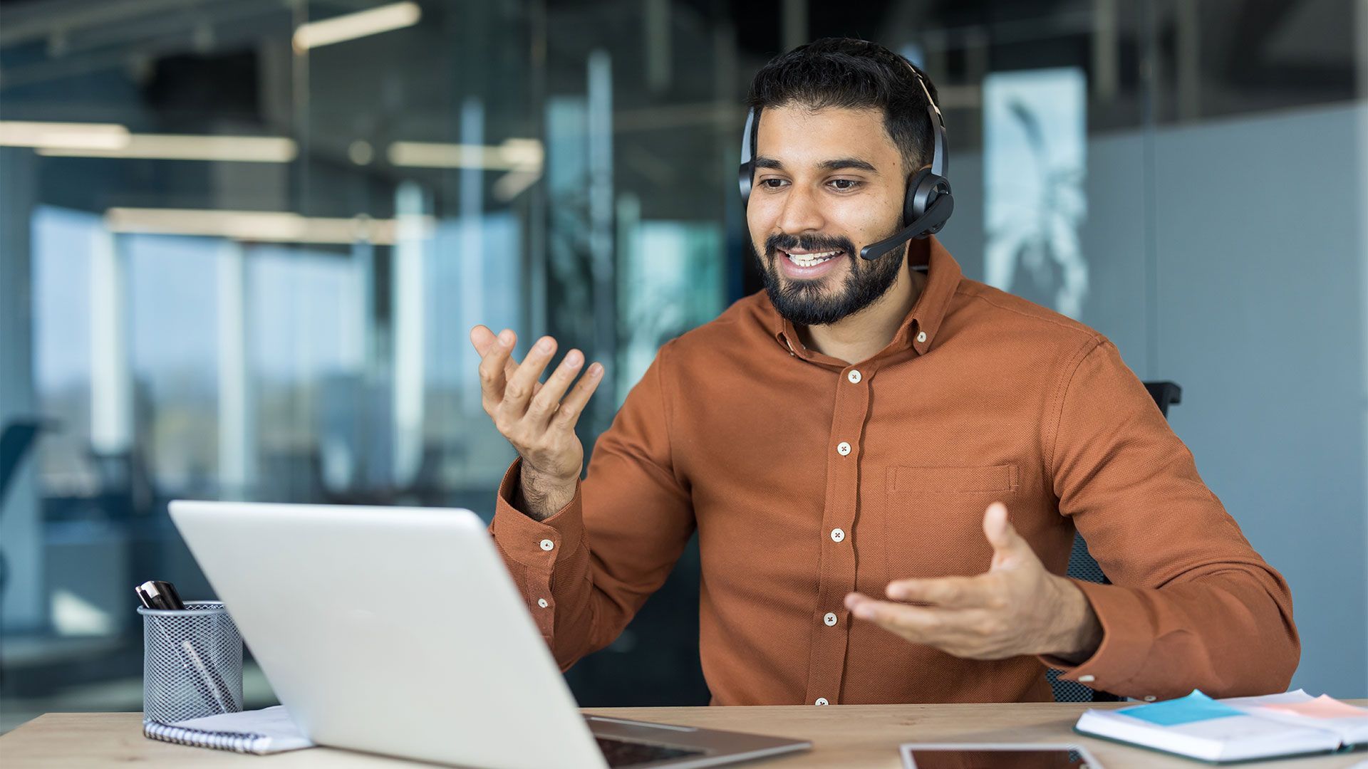 A person wearing a headset and brown shirt sits at a desk, gesturing while having a video call on a laptop.