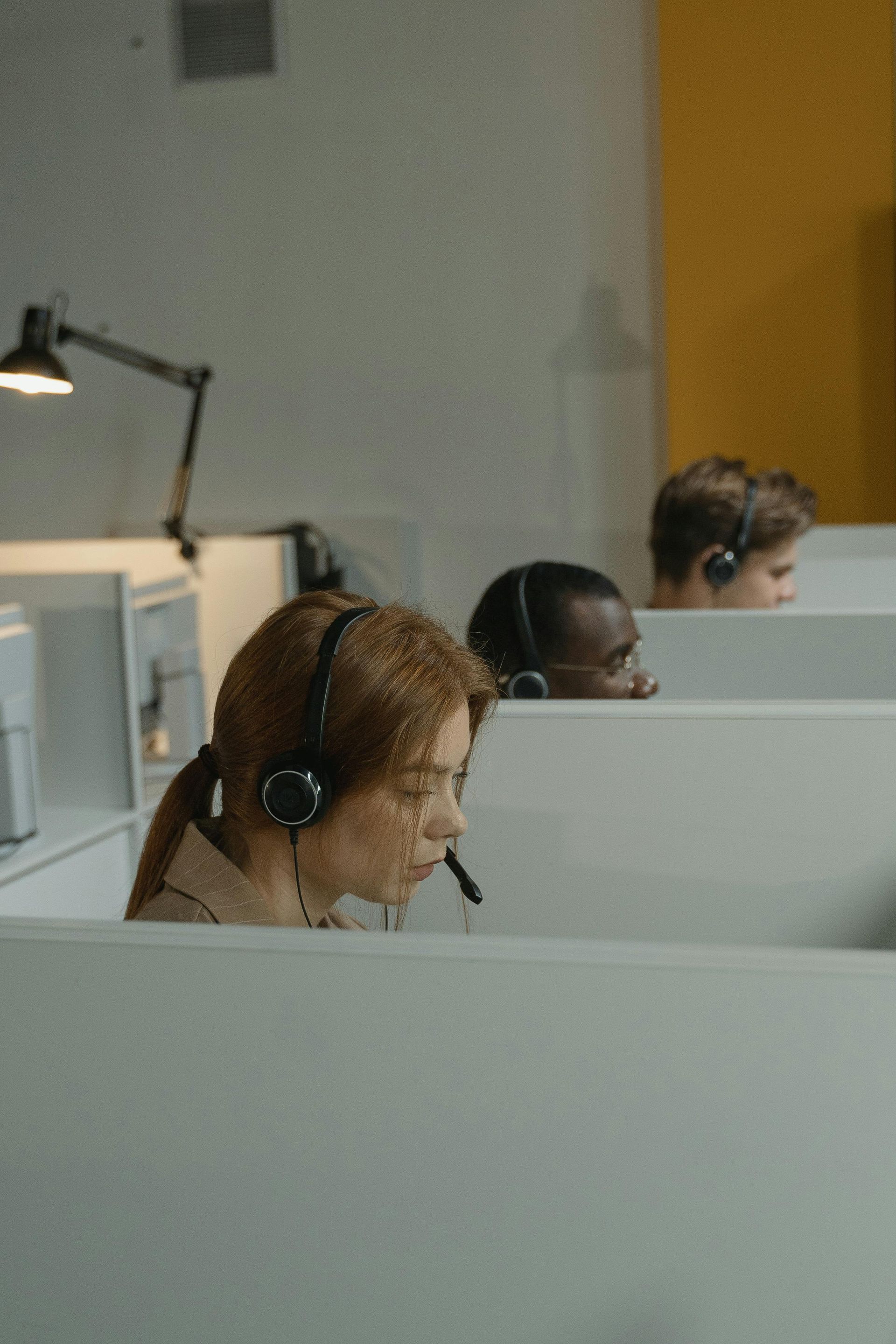 People wearing headsets work in a call center, sitting at desks separated by partitions.