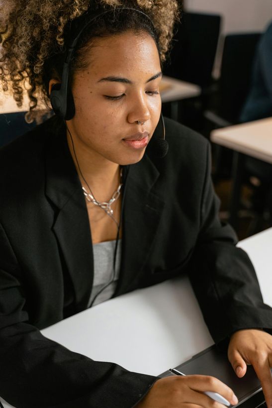 A person in a black blazer and headset writes on a digital tablet at a desk, looking down in concentration.