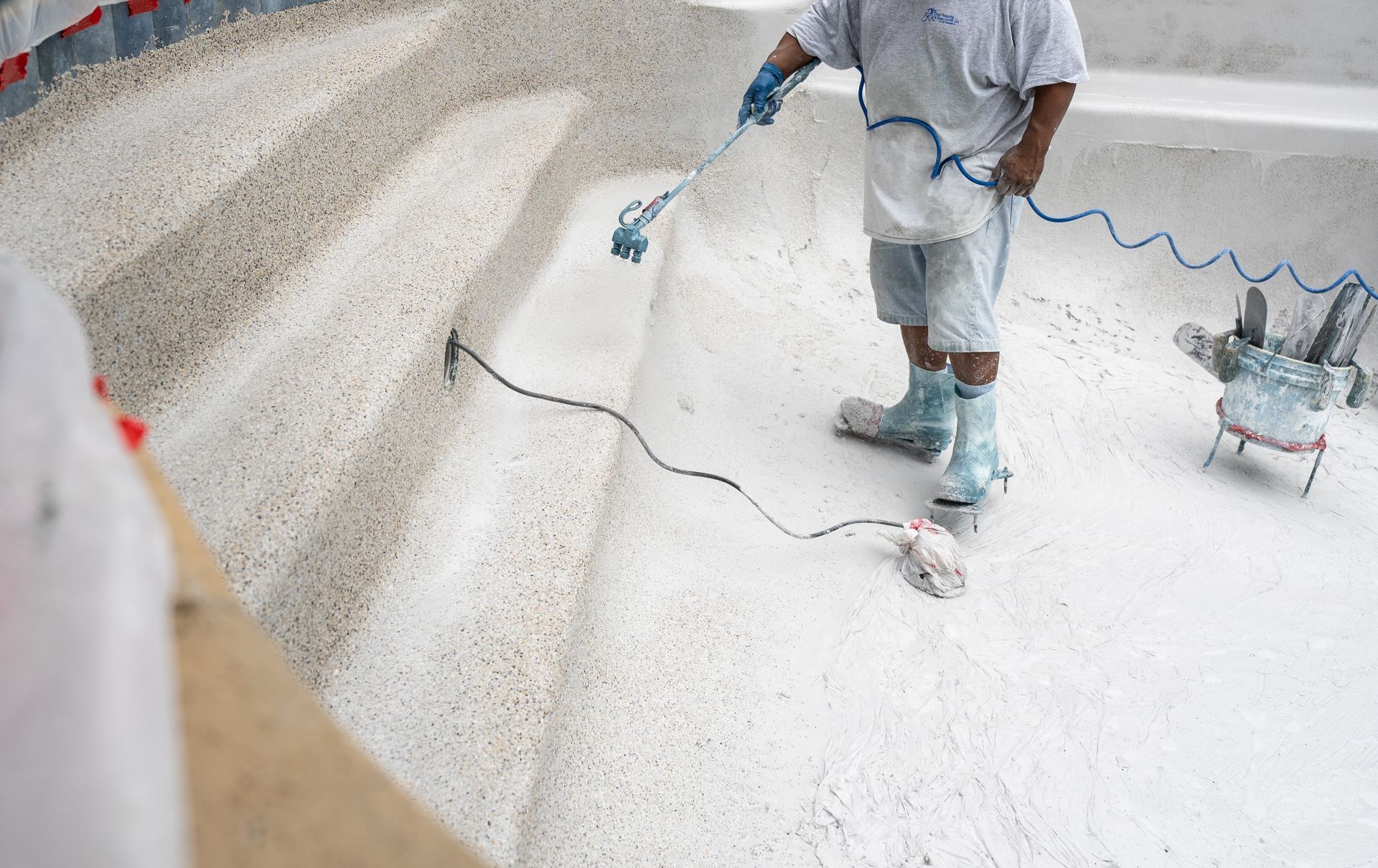 A worker in protective boots resurfaces the interior of a concrete swimming pool using a handheld power tool.