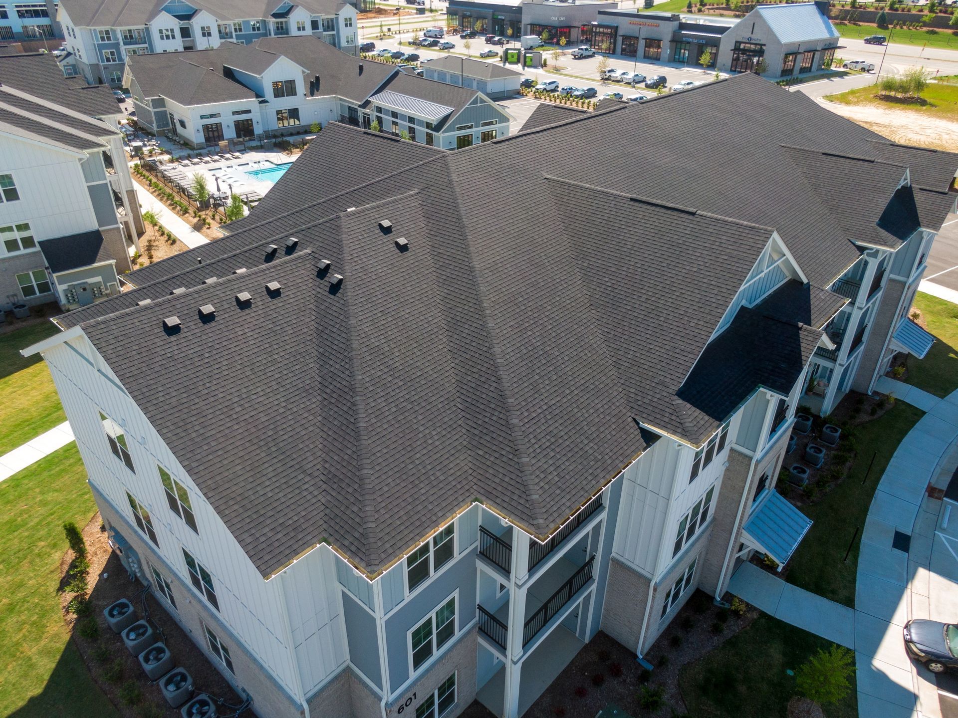 An aerial view of a multi-story apartment building with a dark shingled roof, surrounded by paved areas and green lawns.