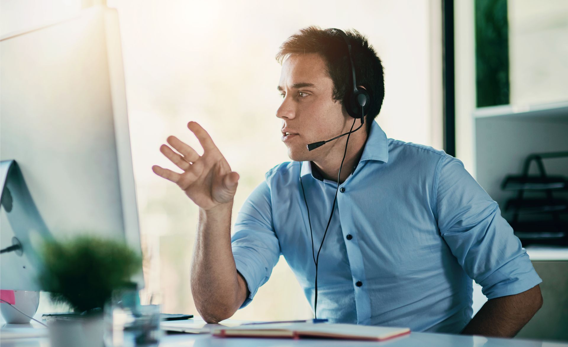 A person in a light blue shirt wearing a headset gestures while speaking to their computer screen in an office.