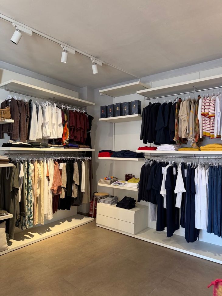 Interno di una boutique di abbigliamento con appendiabiti di capi neutri e scuri su scaffali bianchi e tavoli espositivi.