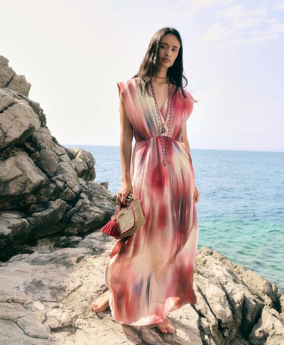 Una donna con un abito rosa è in piedi su una scogliera rocciosa in riva all'oceano, con in mano una borsa di paglia.