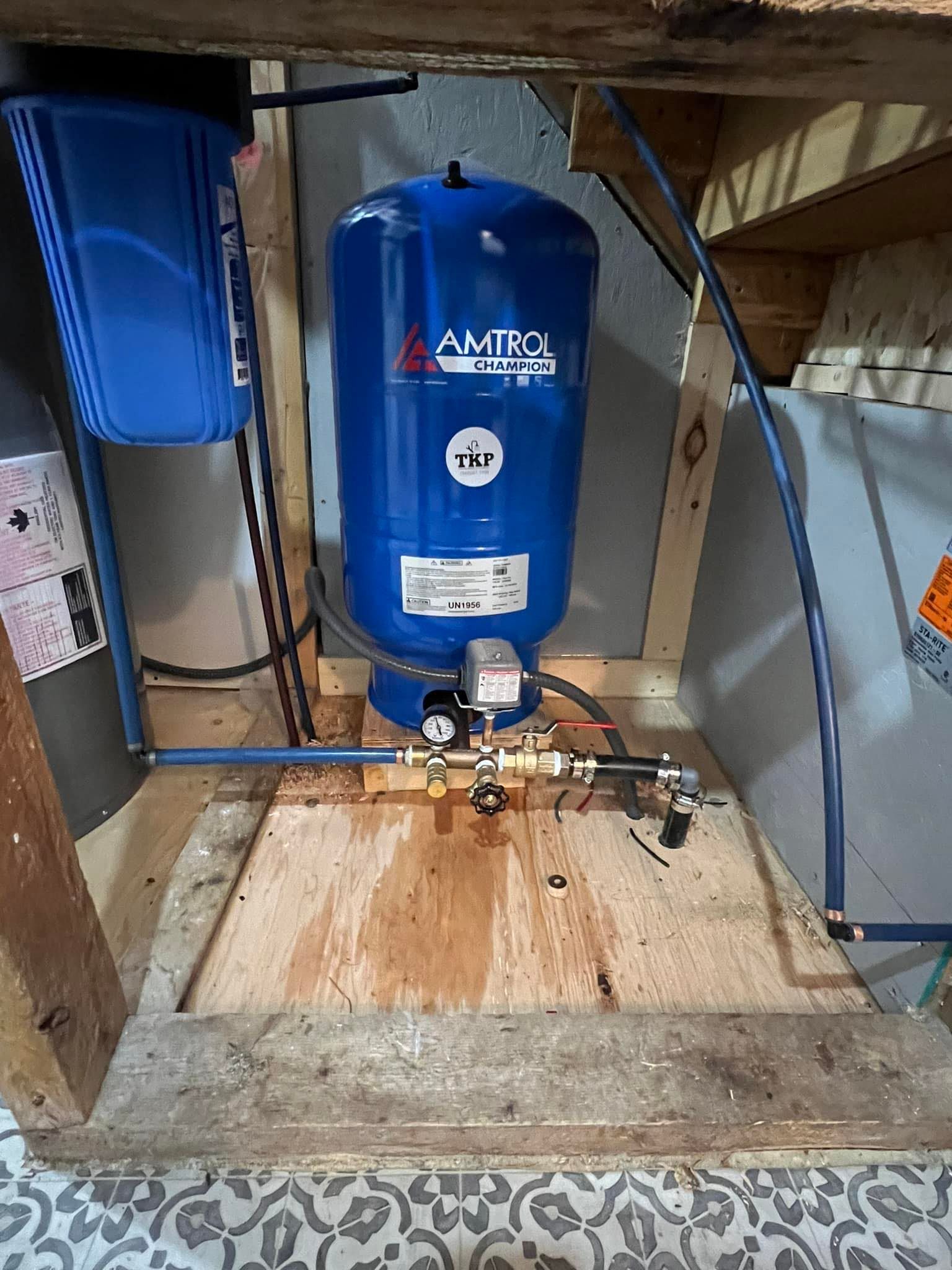A blue water pressure tank and filter system in a wooden enclosure with visible plumbing. Some water is spilled on the wooden platform.