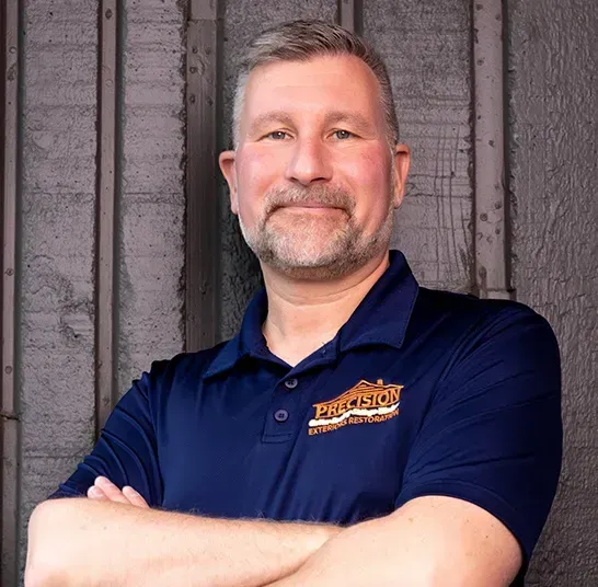 A man with a beard is standing with his arms crossed in front of a wooden wall.