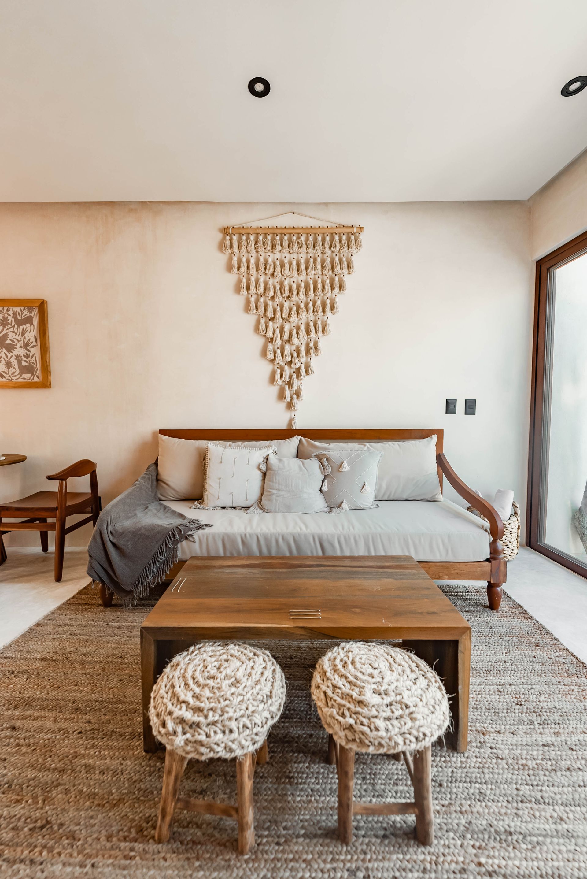 A daybed with cushions and two woven stools sits on a textured rug in a bright, minimalist room with wall decor.