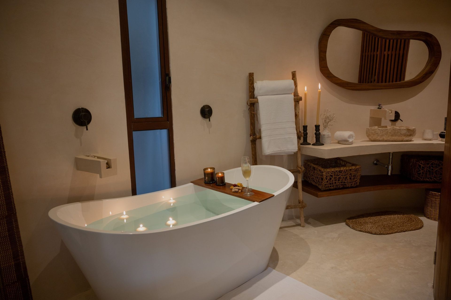 A modern, neutral-toned bathroom with a white soaking tub, a wooden bath tray, a ladder towel rack, and a vanity mirror.
