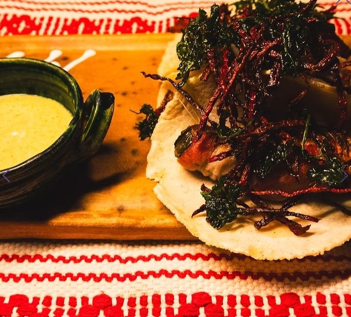 Taco on a wooden board with green sauce, topped with dark greens, on a red-and-white cloth
