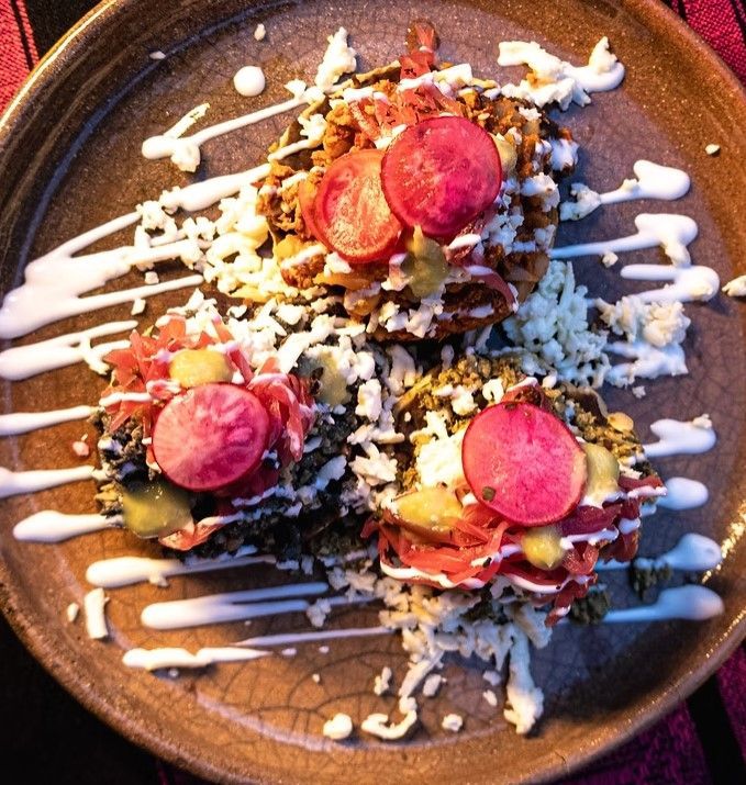 Overhead view of three colorful tacos with pink radish slices and white drizzle on a dark plate