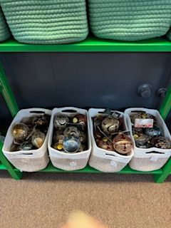 Four white baskets on a shelf, filled with spherical objects, below two green woven baskets.