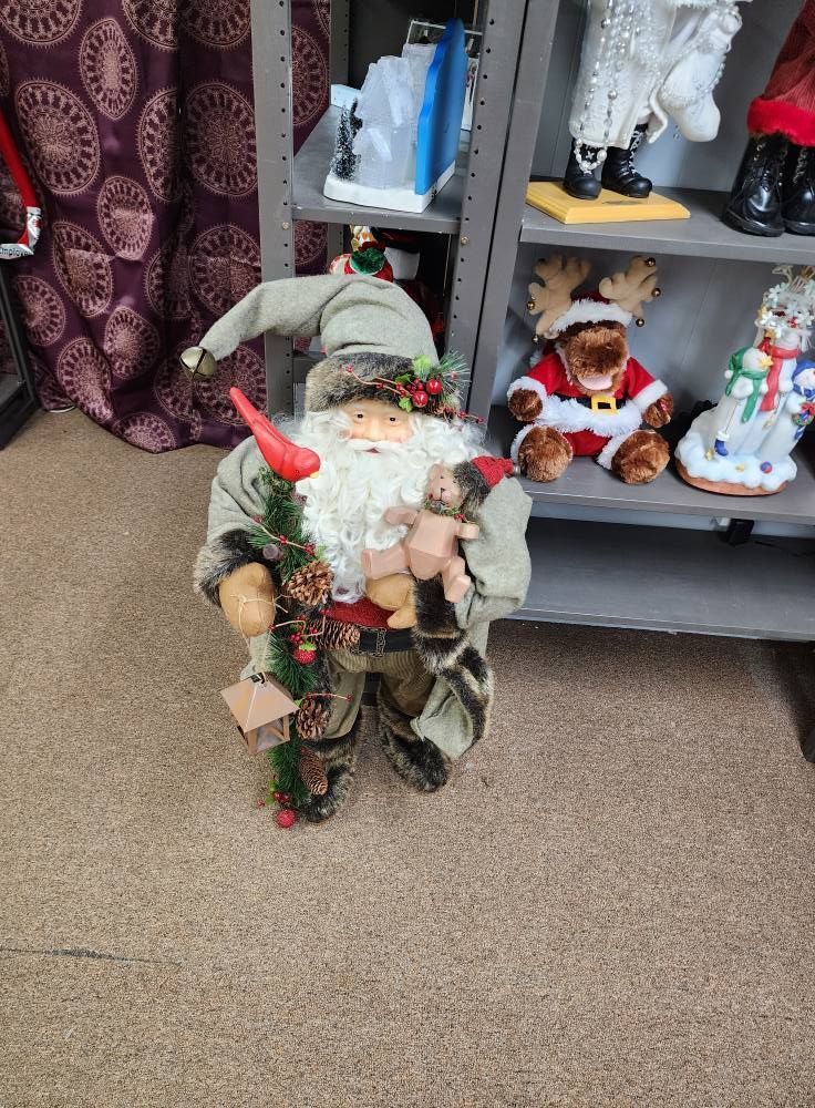 Santa Claus figurine with long hat, holding a doll and a tree, next to shelves with decorations.