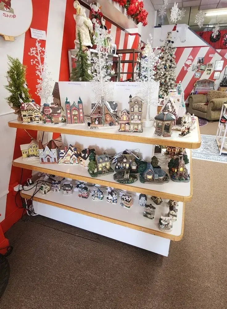 Three-tiered shelf displaying miniature lighted Christmas village houses and figurines in a shop setting.
