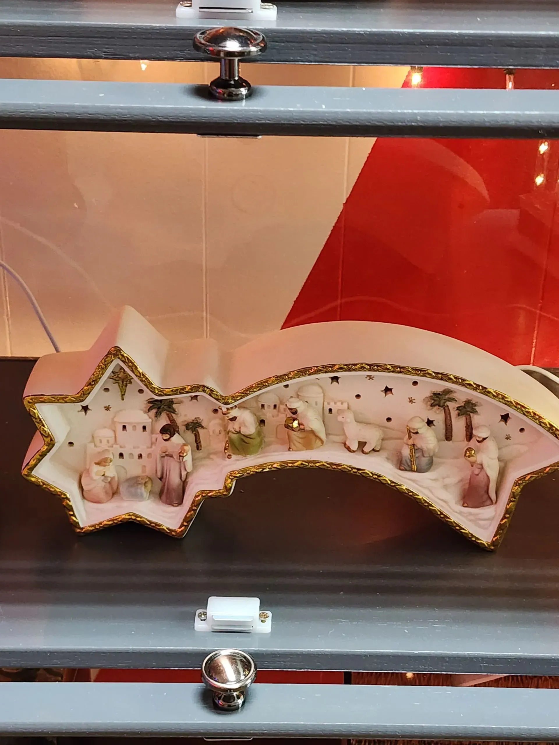 Nativity scene inside a star-shaped ornament with gold trim, housed in a glass display case with red and white background.