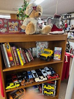 Wooden bookshelf filled with toys, books, and model cars. Top shelf: teddy bear, elephant, book. Middle: books, toy truck, card. Bottom: model cars.