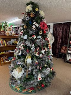 Christmas tree decorated with ornaments, tinsel, and a tree skirt in a room with shelves and curtains.