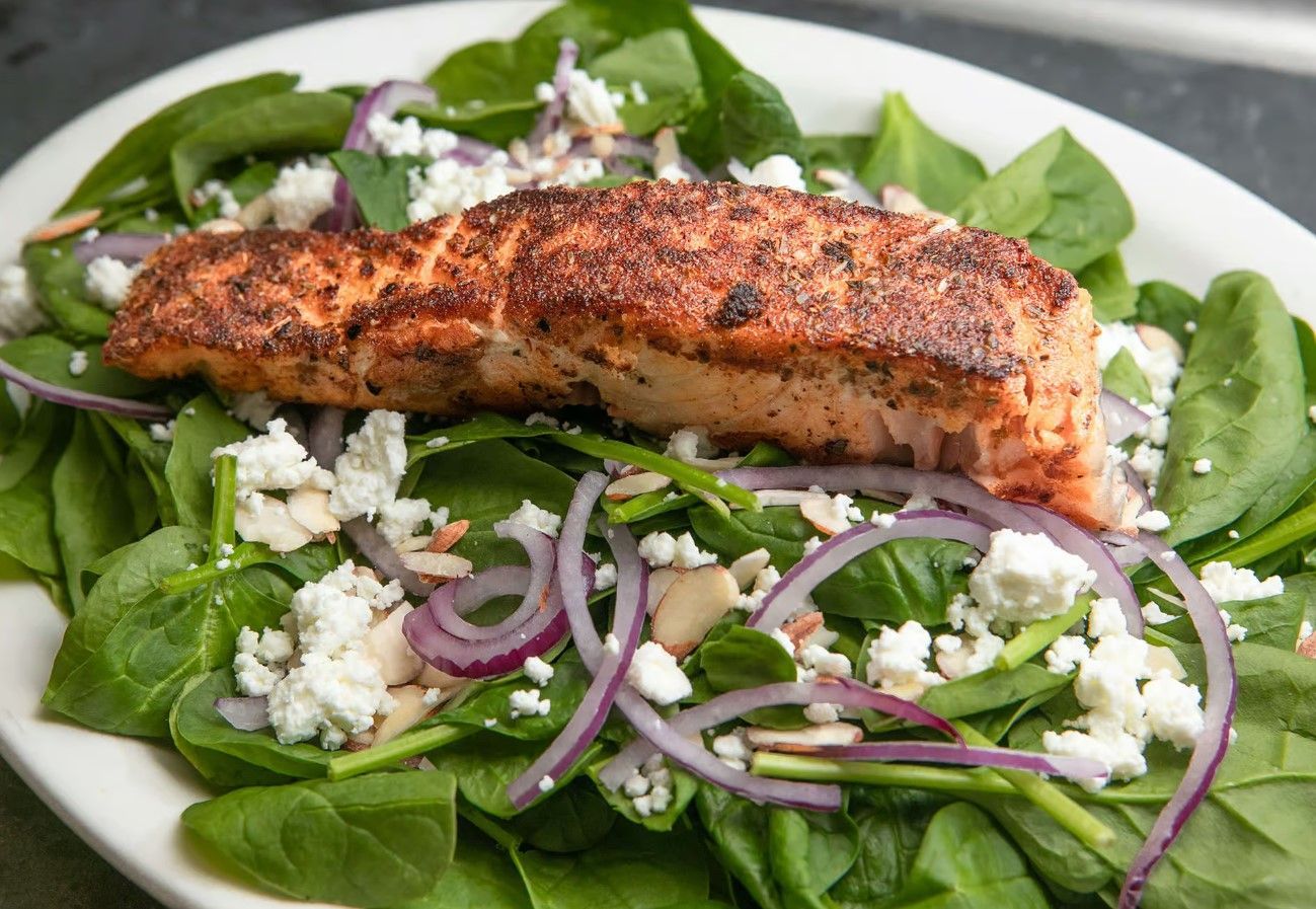 A white plate with blackened salmon over a bed of mixed greens with red onion, sliced almonds and feta cheese