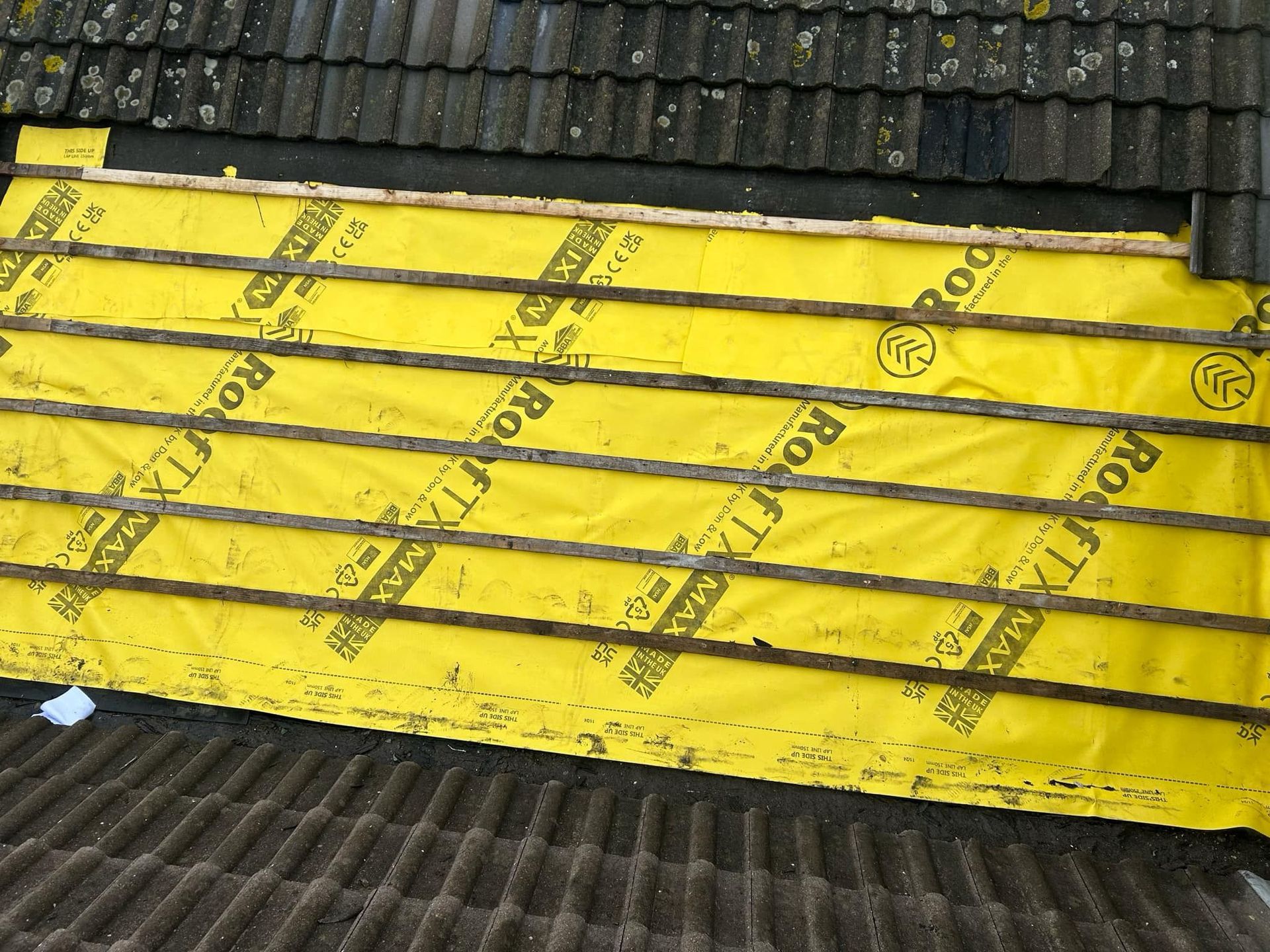 Roofer kneeling on a roof, installing tiles. A stack of tiles and a chimney are in the background.