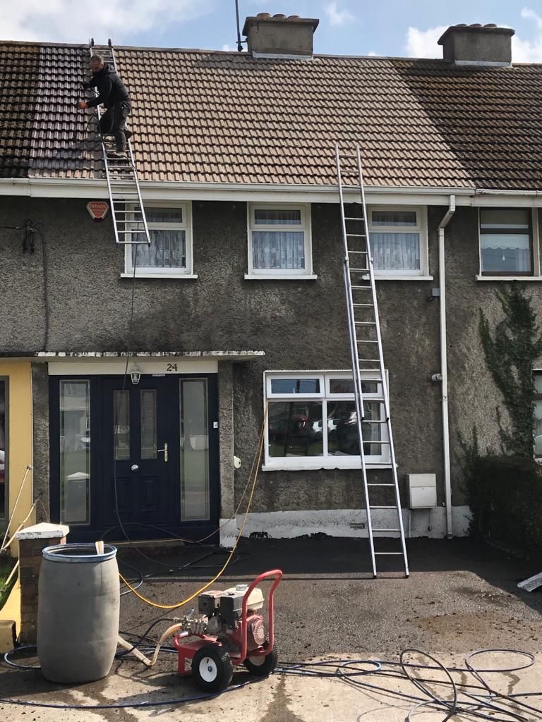 Person on a ladder power washing a tiled roof of a house; a pressure washer sits below.