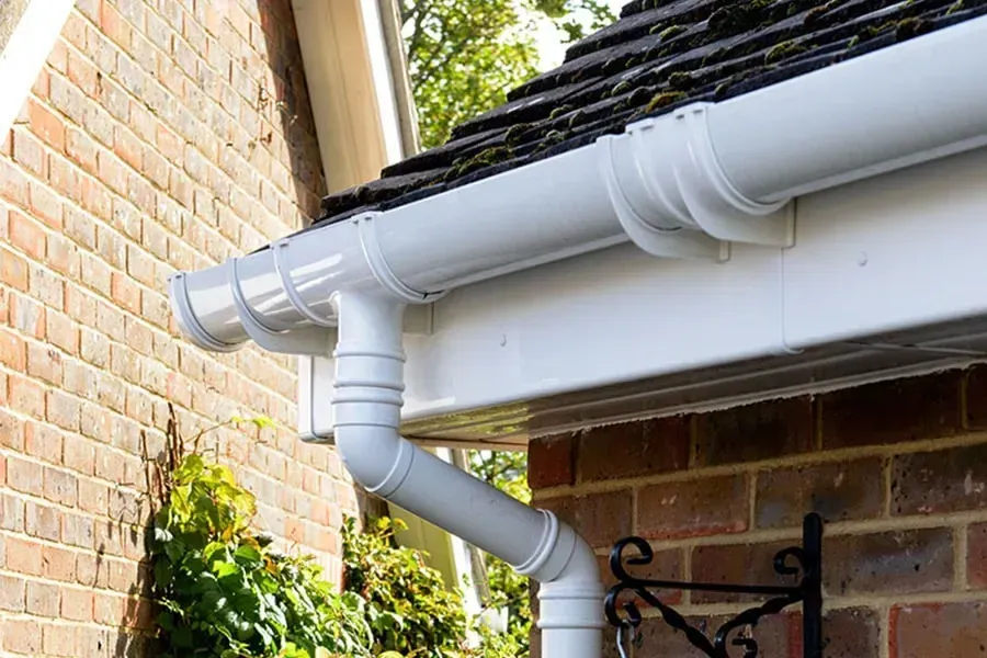 White gutter system on a brick house with a dark roof and green foliage.