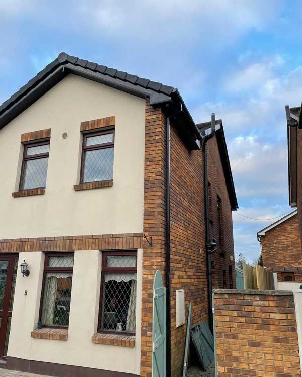Two-story house with cream stucco and brick facade, brown window frames, and black roof against a cloudy sky.