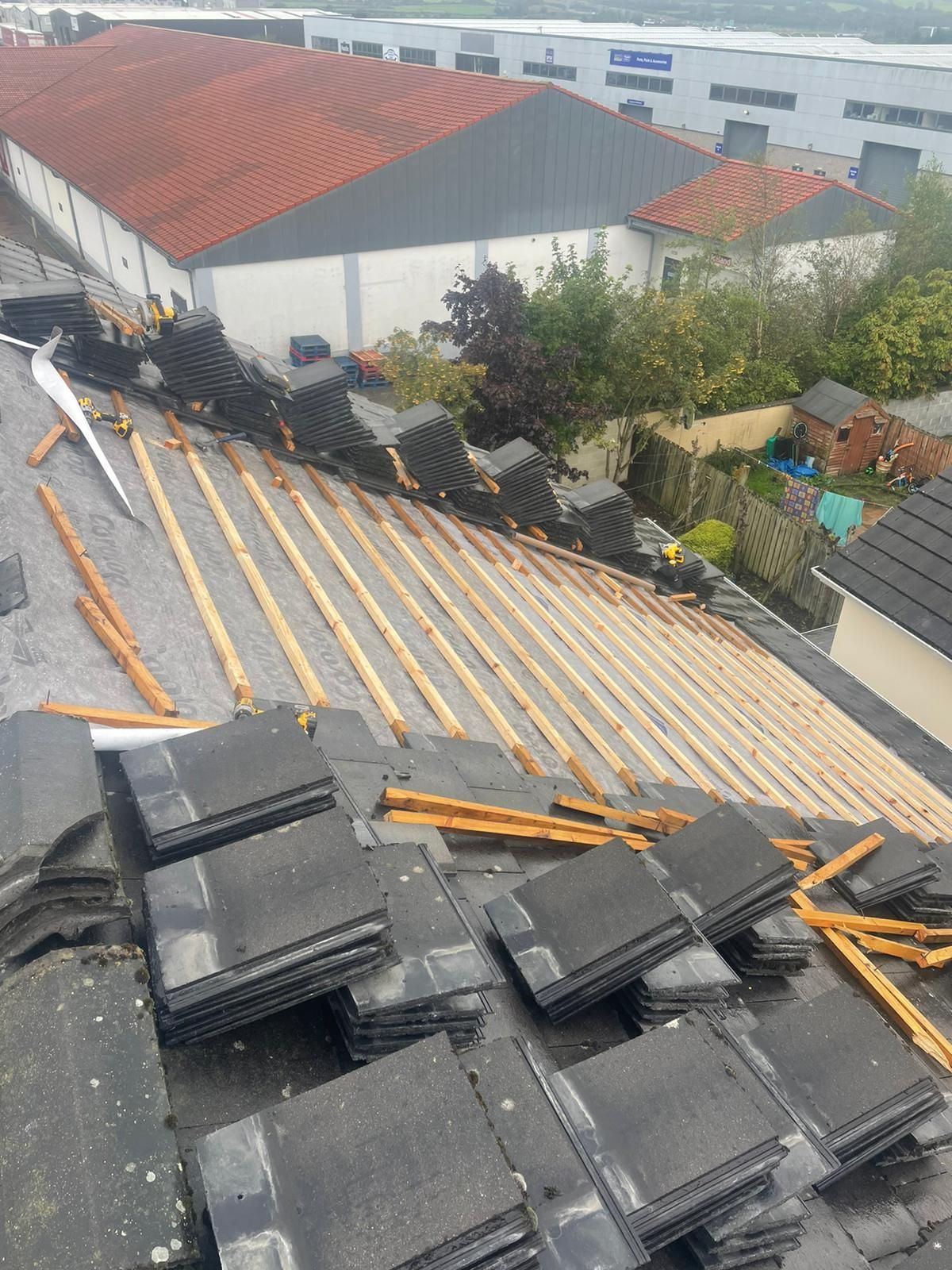 Rooftop partially dismantled, showing exposed wooden supports and stacks of gray tiles; other buildings in background.