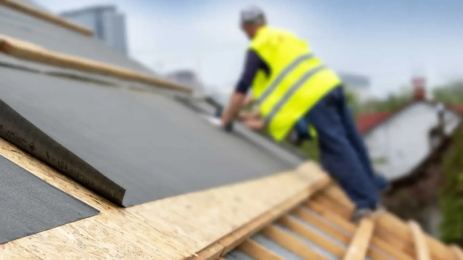 Roof under construction with blue underlayment, wooden beams, and stacked dark tiles.