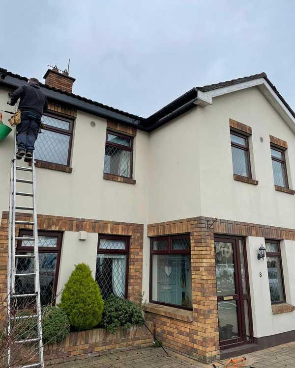 Person on ladder cleaning gutters of a two-story beige house with brown trim and brick accents; overcast sky.