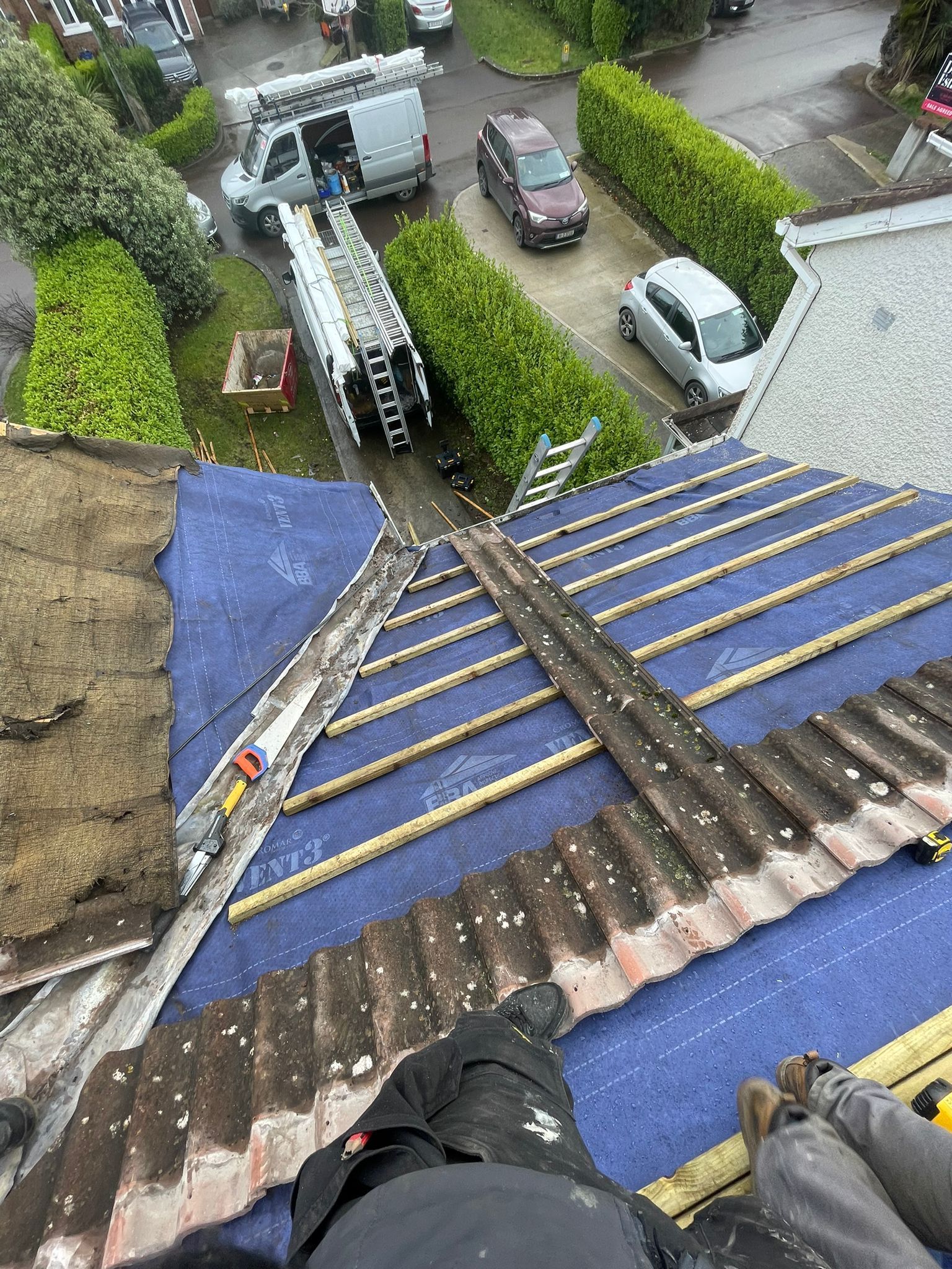 Roofer kneeling on a roof, installing dark gray tiles, wearing safety harness and gloves, in a residential area.