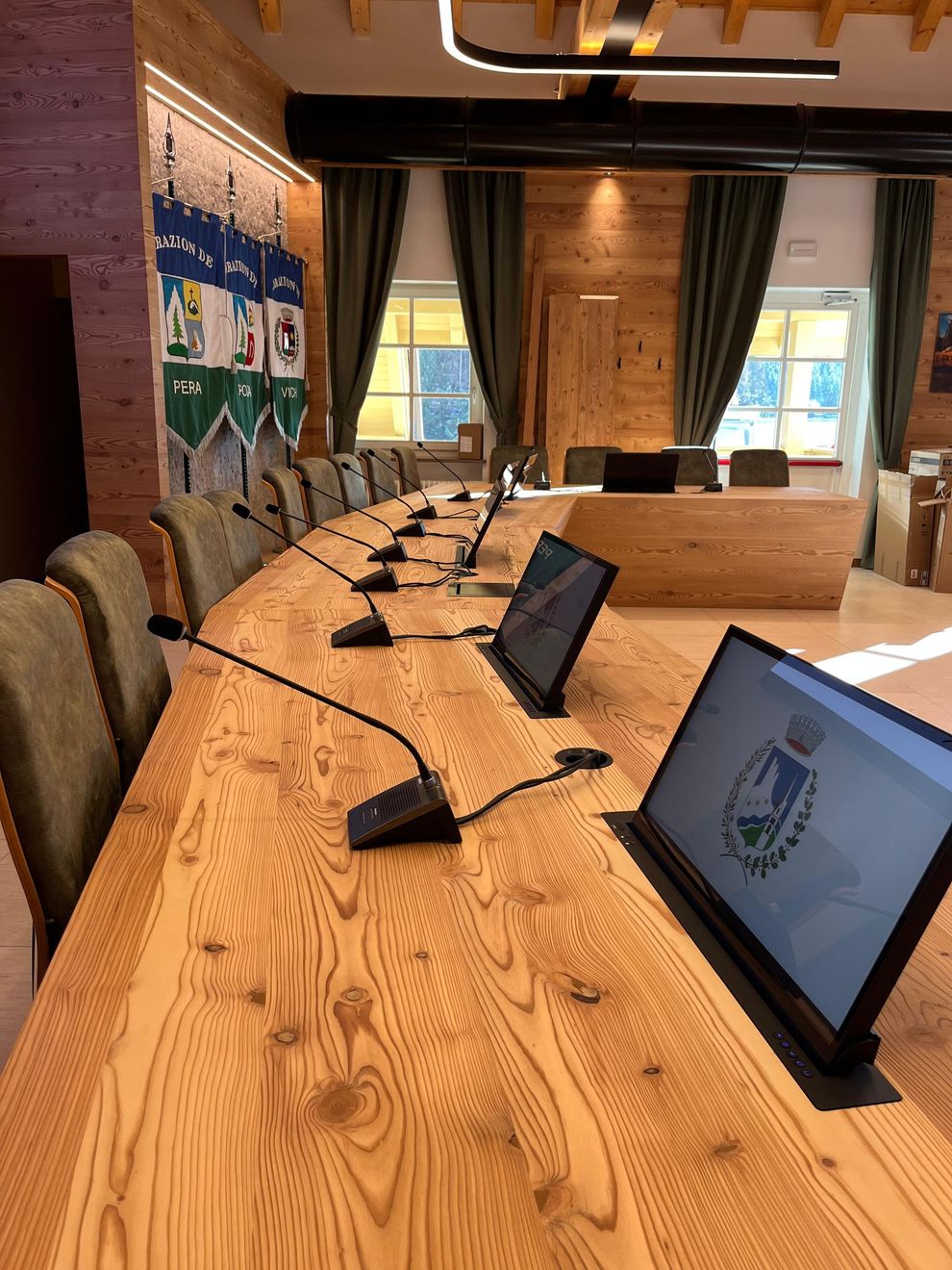 Sala conferenze con tavolo in legno curvo, computer portatili e microfoni di fronte a una parete finestrata.