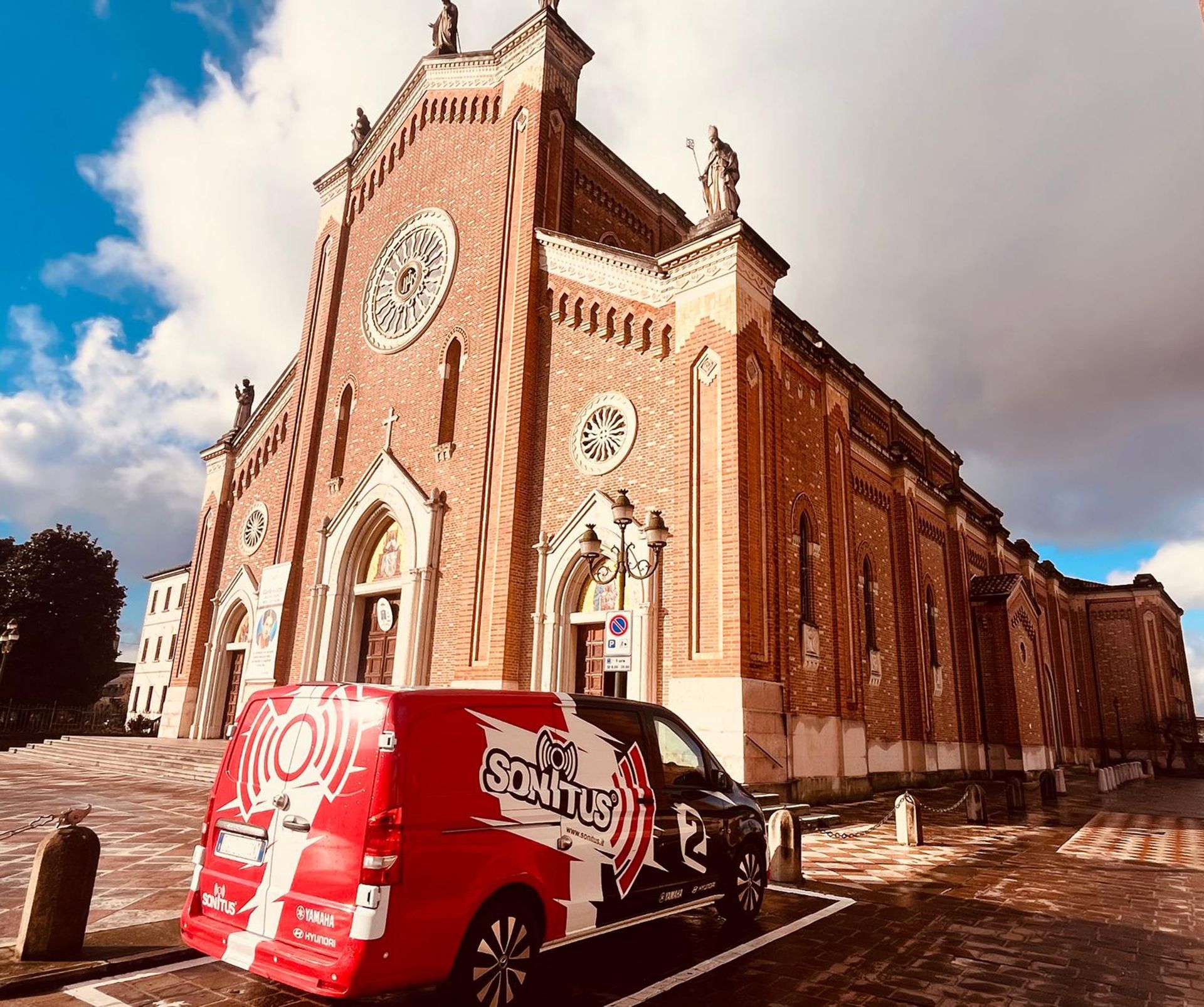 Interno di una grande chiesa con file di banchi, soffitti alti e un altare in fondo. Due paraventi con quadri gialli sono appesi alle pareti.