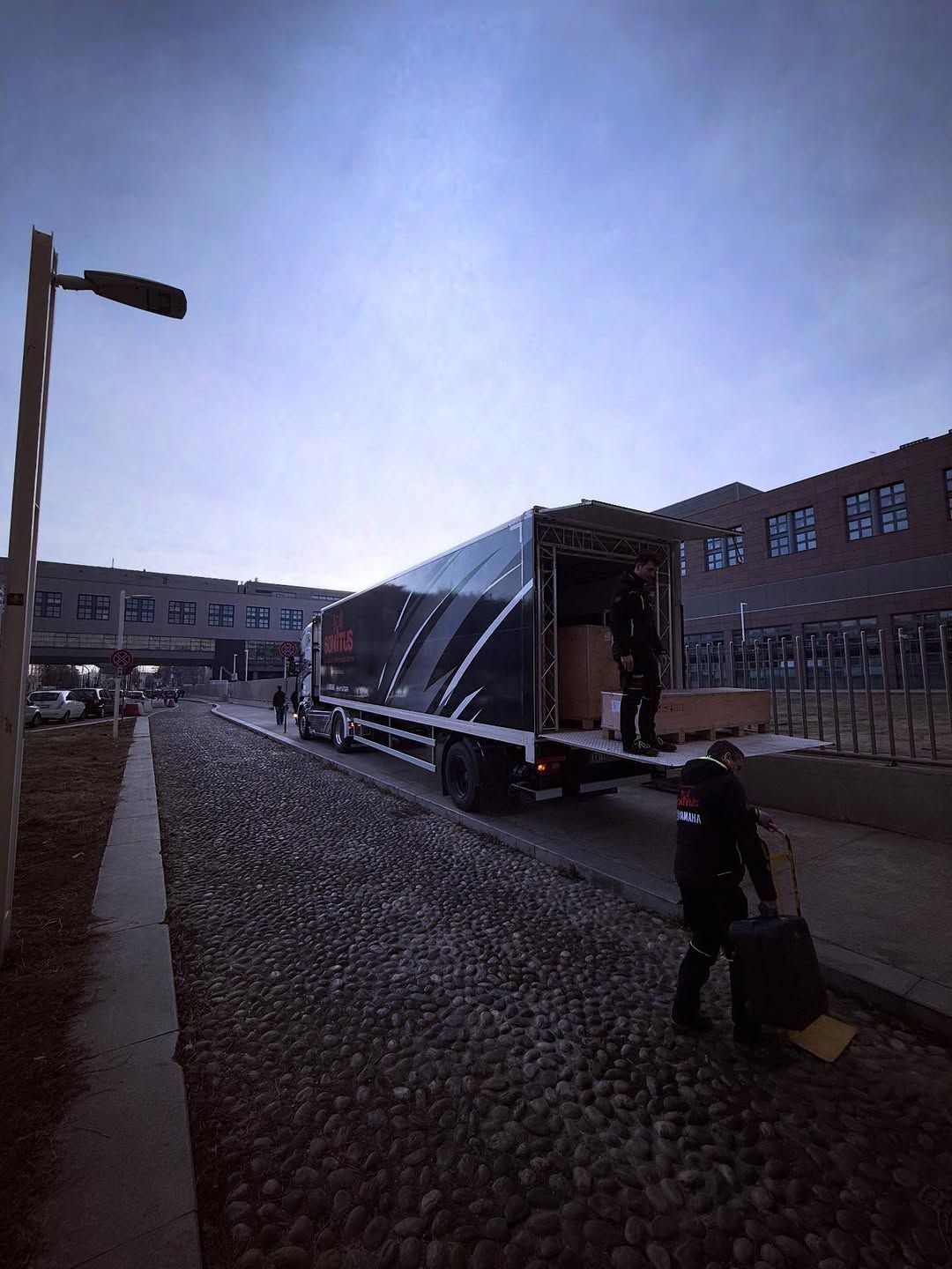 Un camion scarica rifornimenti all'ingresso di un edificio. Una persona trasporta bagagli. Nuvoloso, luce diurna fresca.