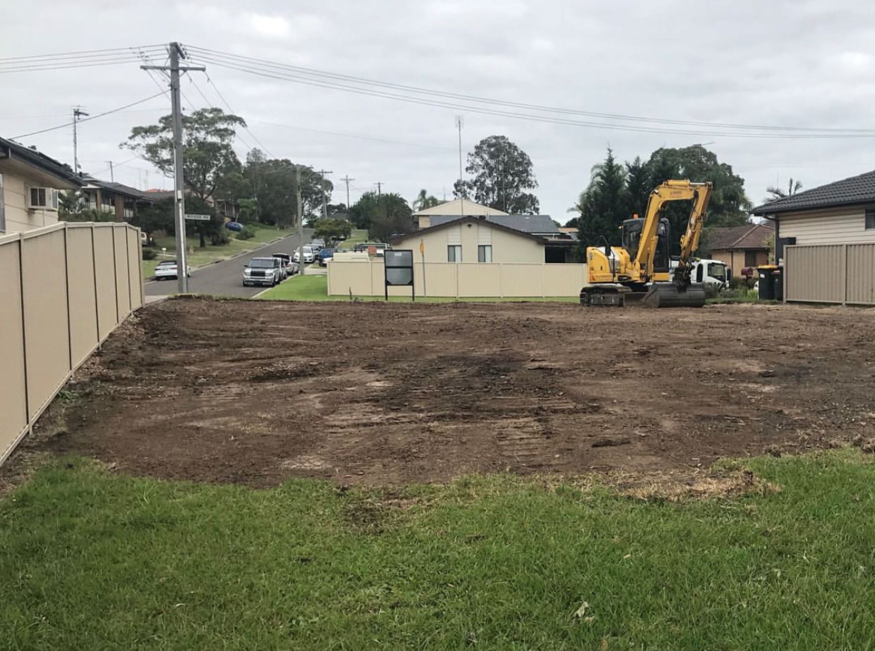 A yellow excavator is working on a dirt field in front of a house.