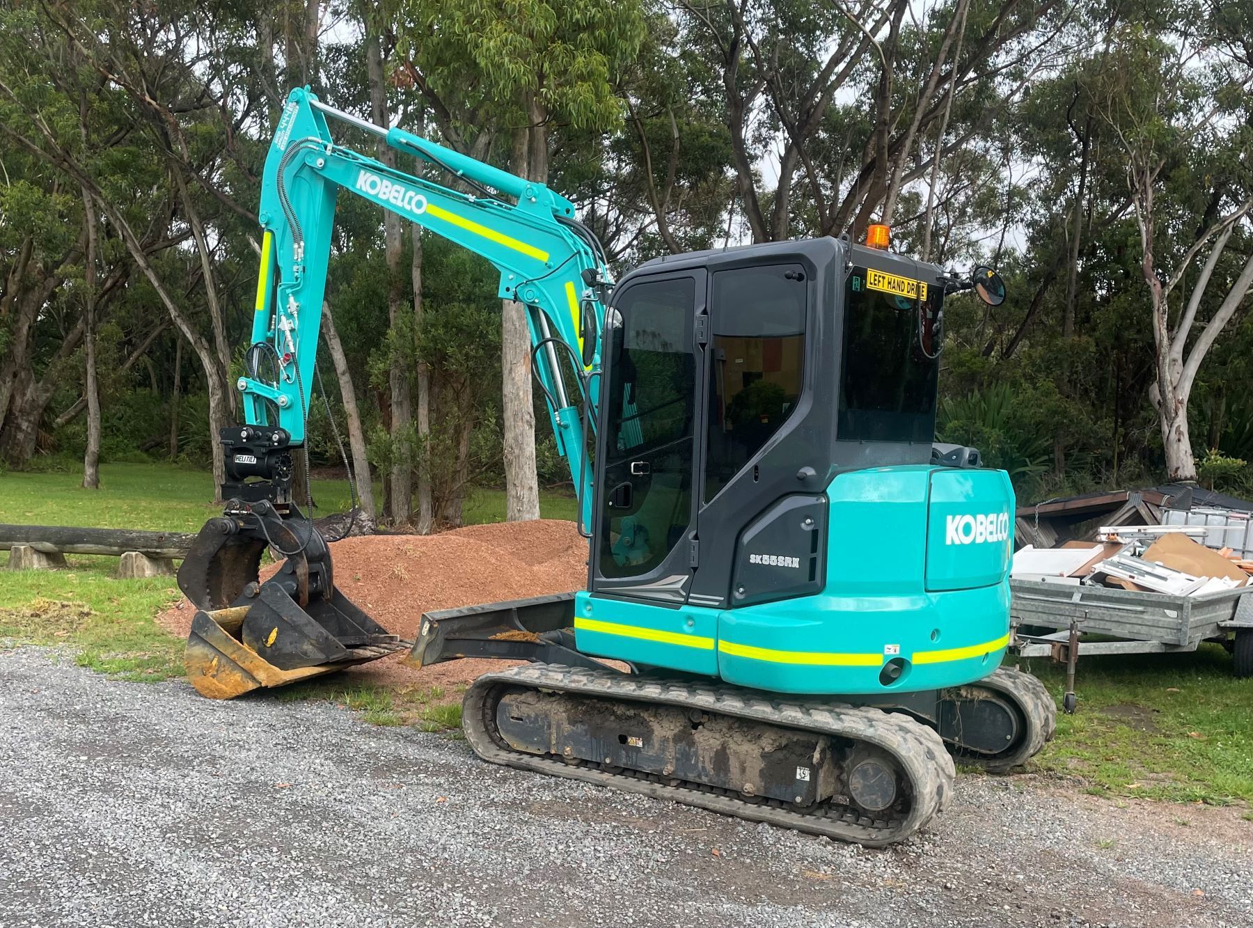 A blue excavator is parked in a gravel lot next to a trailer.