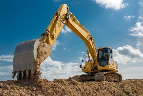 A yellow excavator digging dirt at a construction site under a blue sky with scattered white clouds