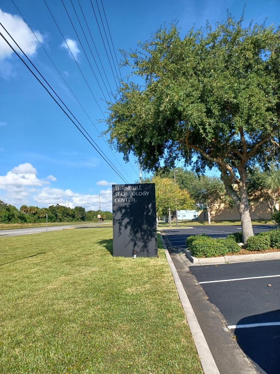 Black sign on a grassy lawn with trees against a blue sky.