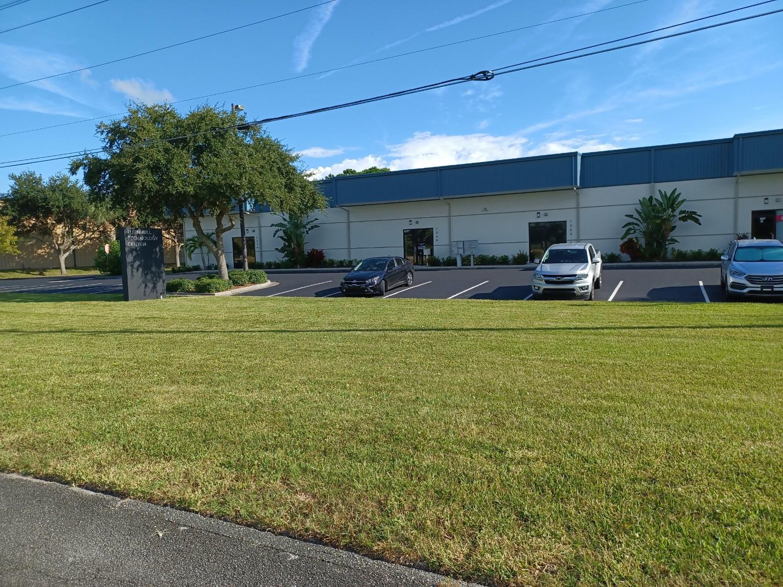 Exterior of a building with parking spaces; green grass in the foreground. Cars parked in spaces. Clear, blue sky.