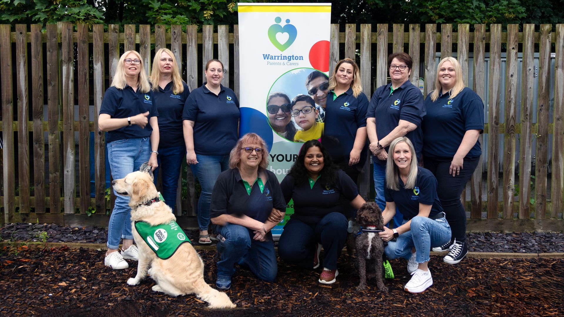 A group of women and a dog are posing for a picture in front of a wooden fence.