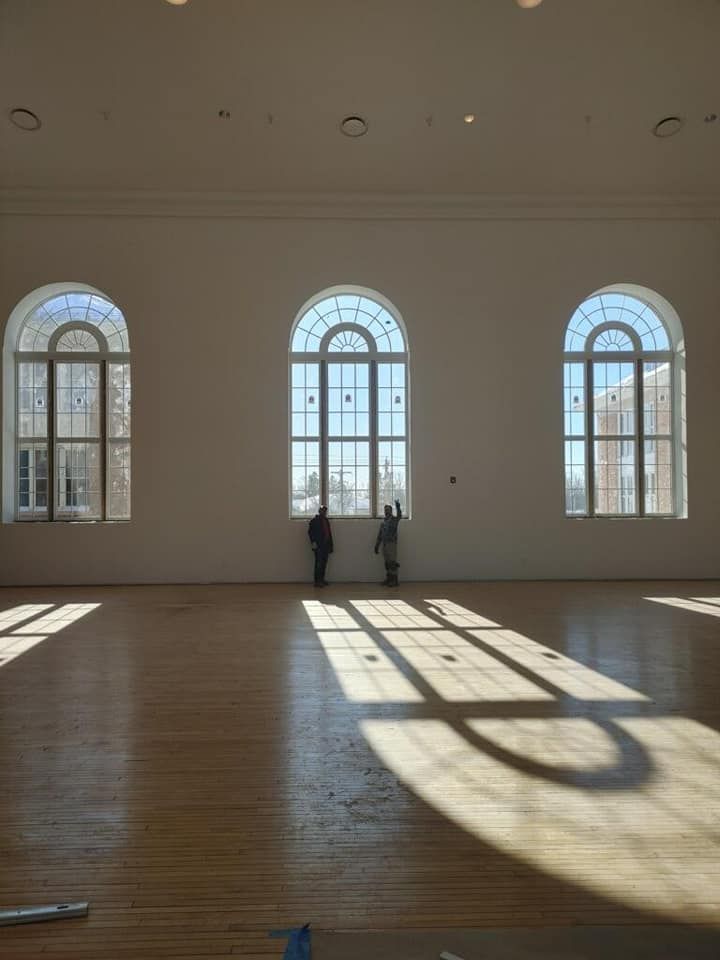 Empty sunlit hall with tall arched windows, long shadows on wooden floor, and two people near the center.