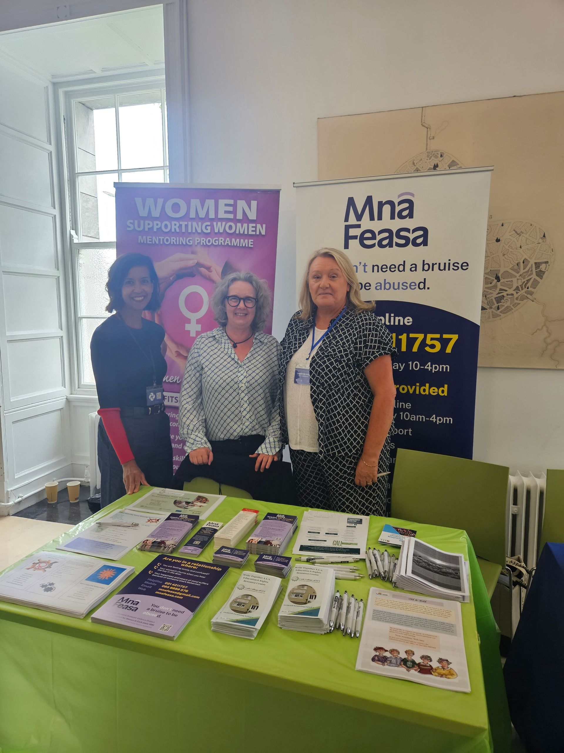 Three women stand behind a table with brochures. Event banner in background.