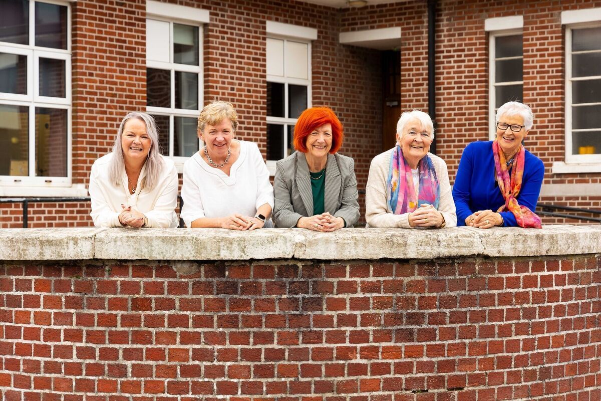 Five women smiling, leaning on a brick wall in front of a brick building.
