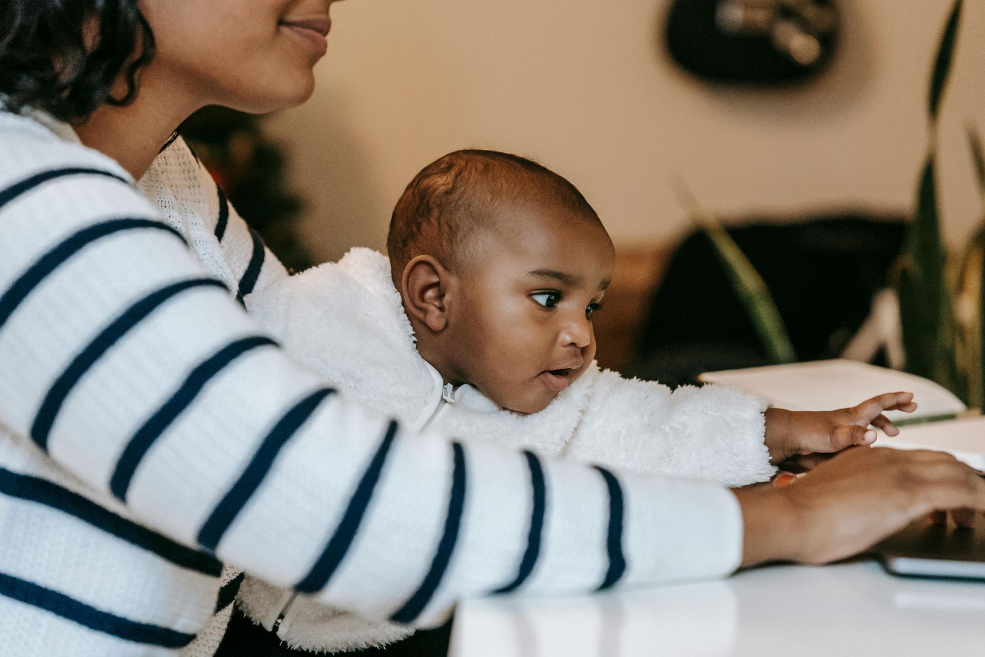 Woman in striped sweater works on a laptop with a baby in a fluffy jacket on her lap.