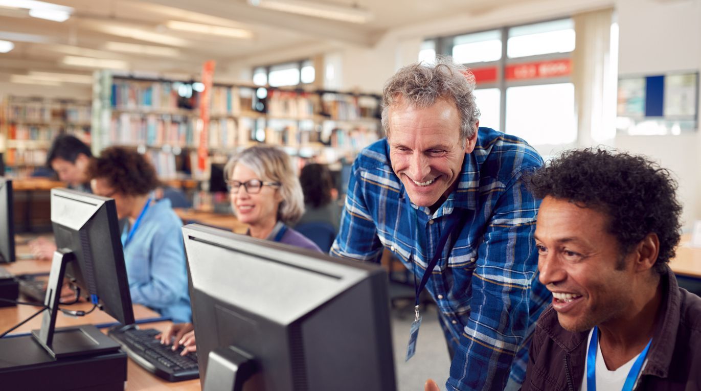 Teacher helping student at a computer; library setting, smiling.