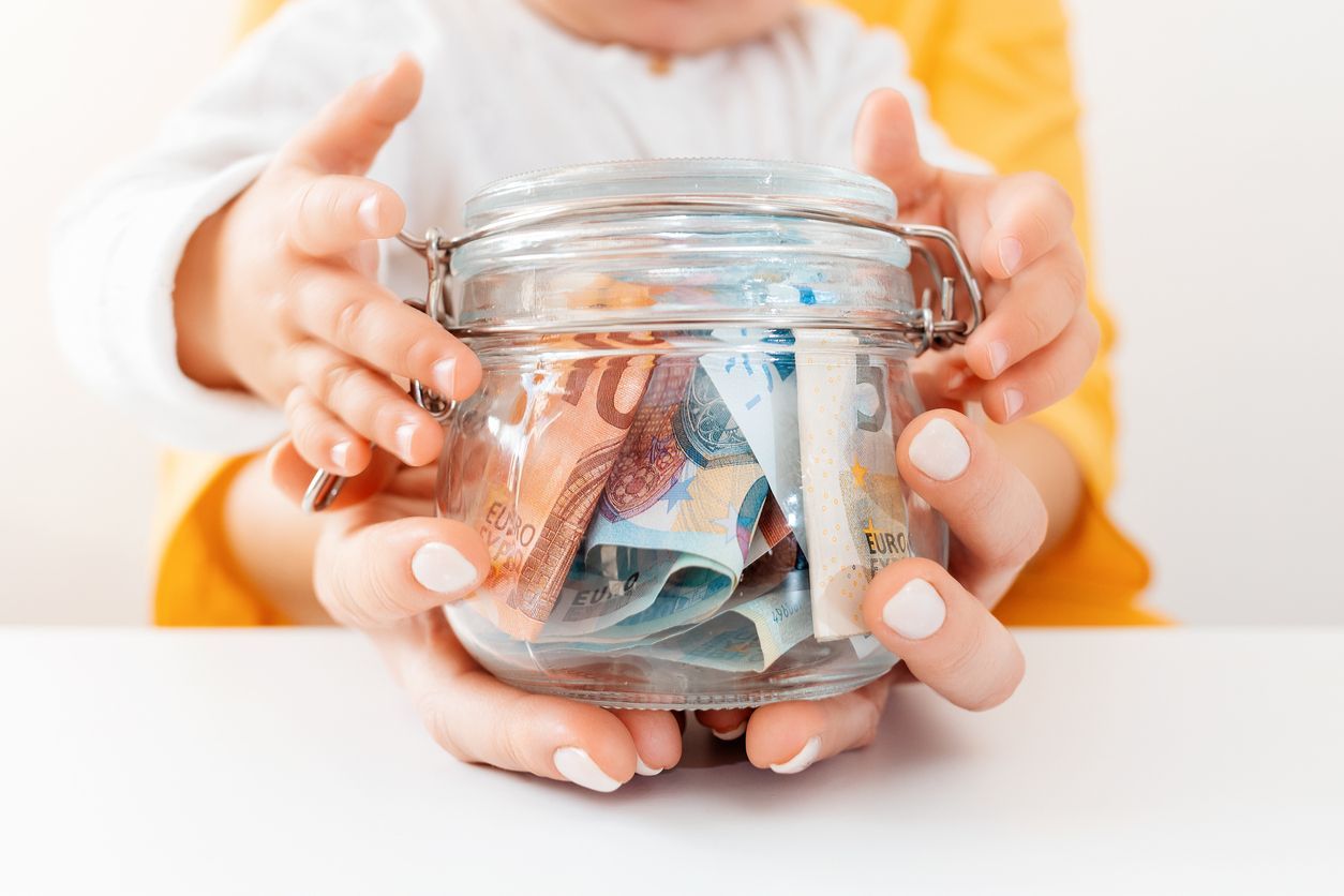 Hands of an adult and child holding a jar filled with money, suggesting savings.