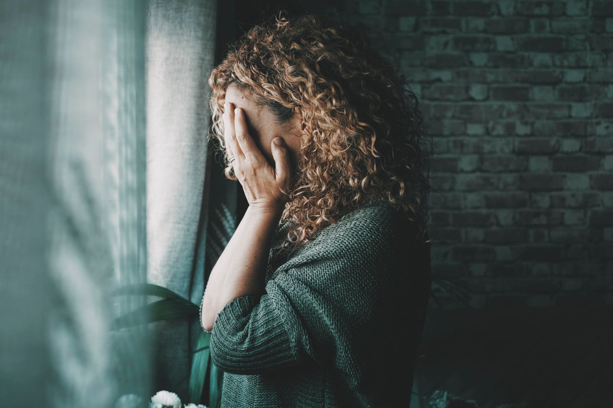 Woman covering face, standing by window, looking distressed. Curly hair, brick wall.
