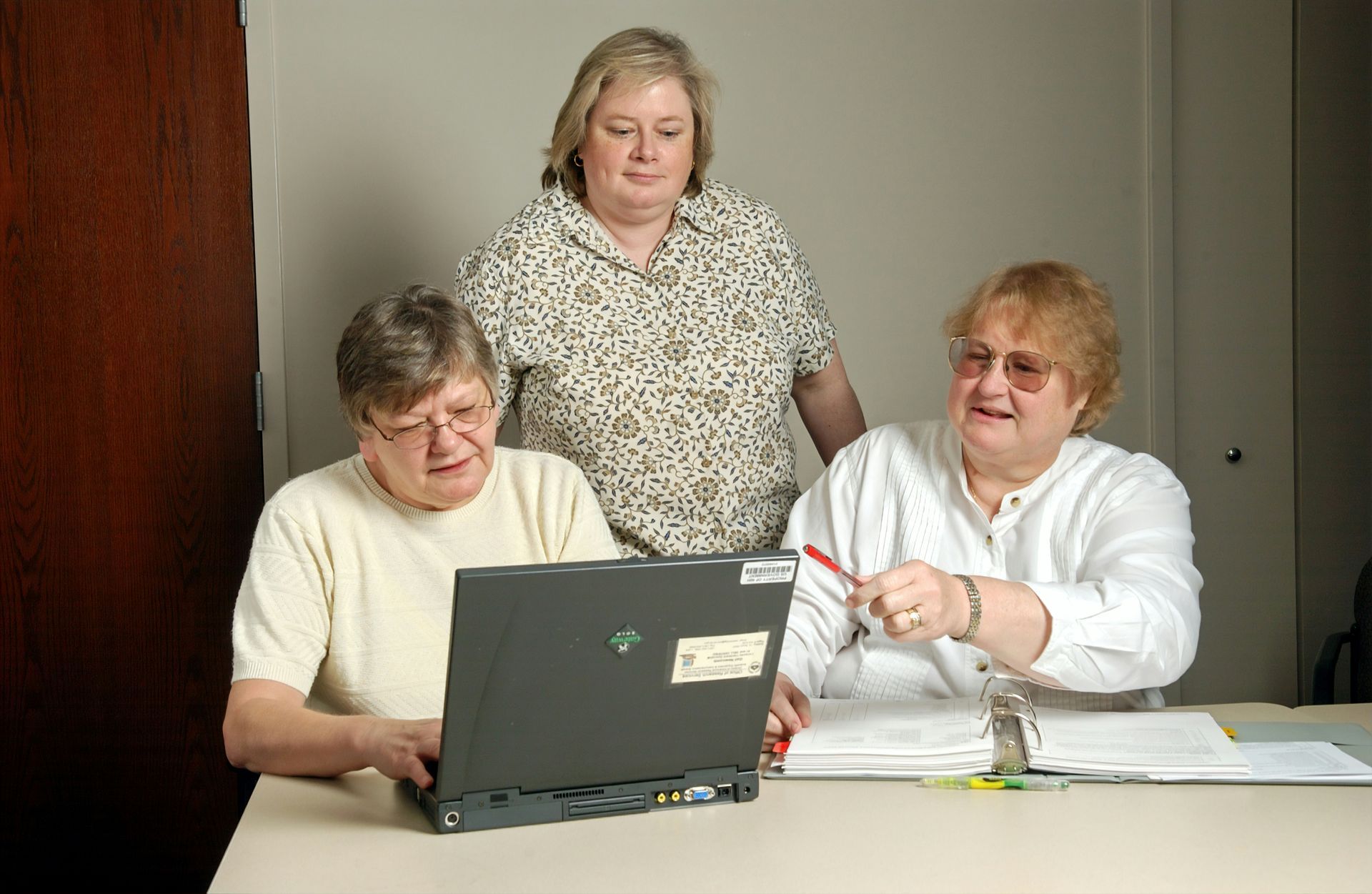 Three people collaborating around a laptop and papers in an office setting. One points a pencil at the screen.