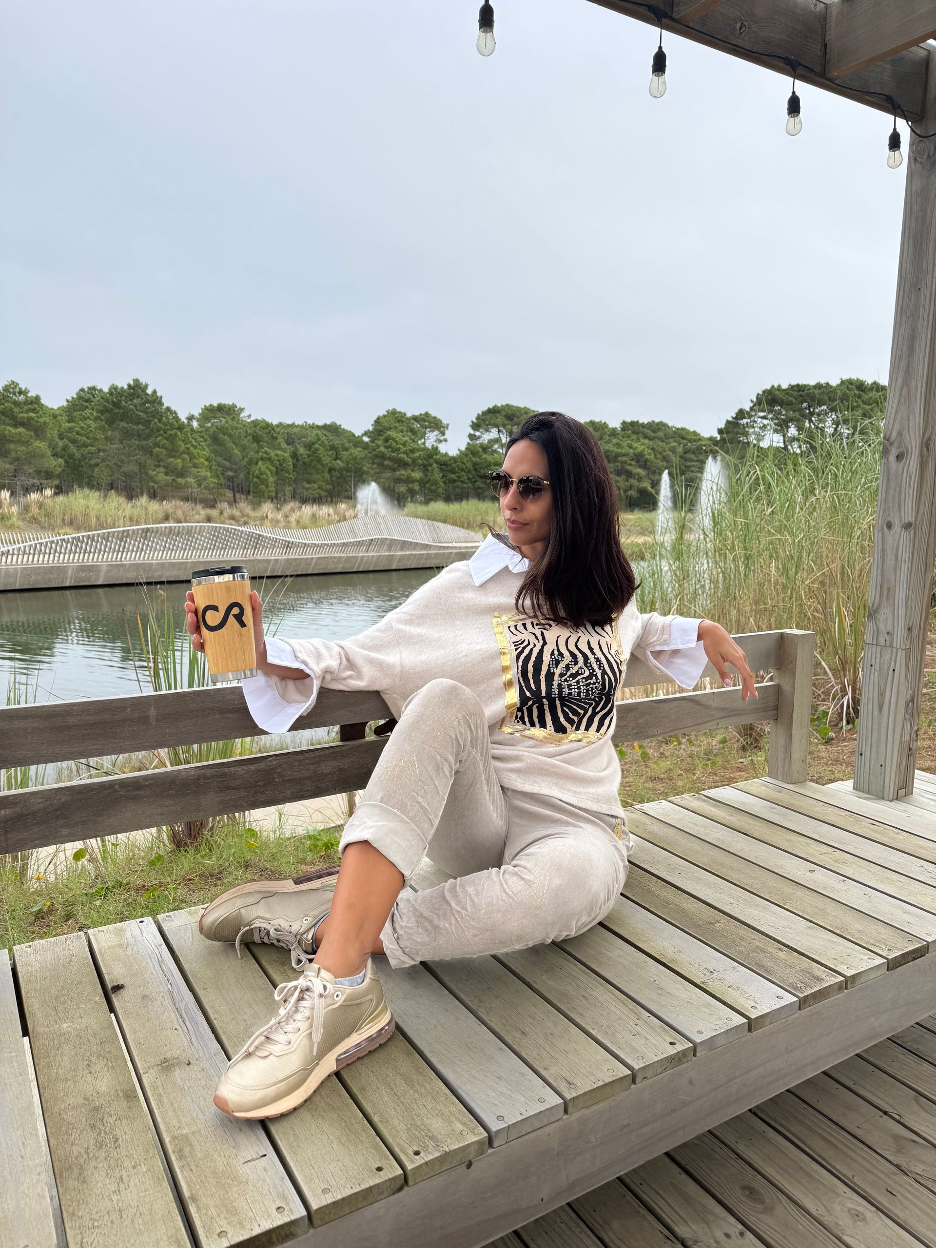 Mujer con gafas de sol sosteniendo una taza de café, sentada en un banco cerca de un estanque con fuentes.