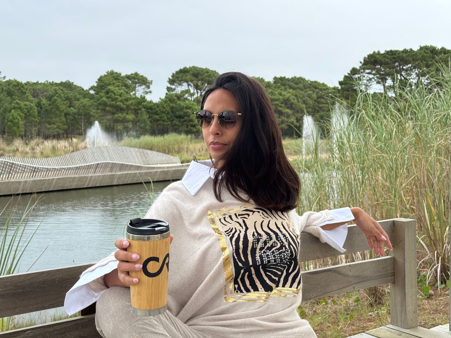 Mujer con gafas de sol sosteniendo una taza de café, sentada en un banco cerca de un estanque con fuentes.