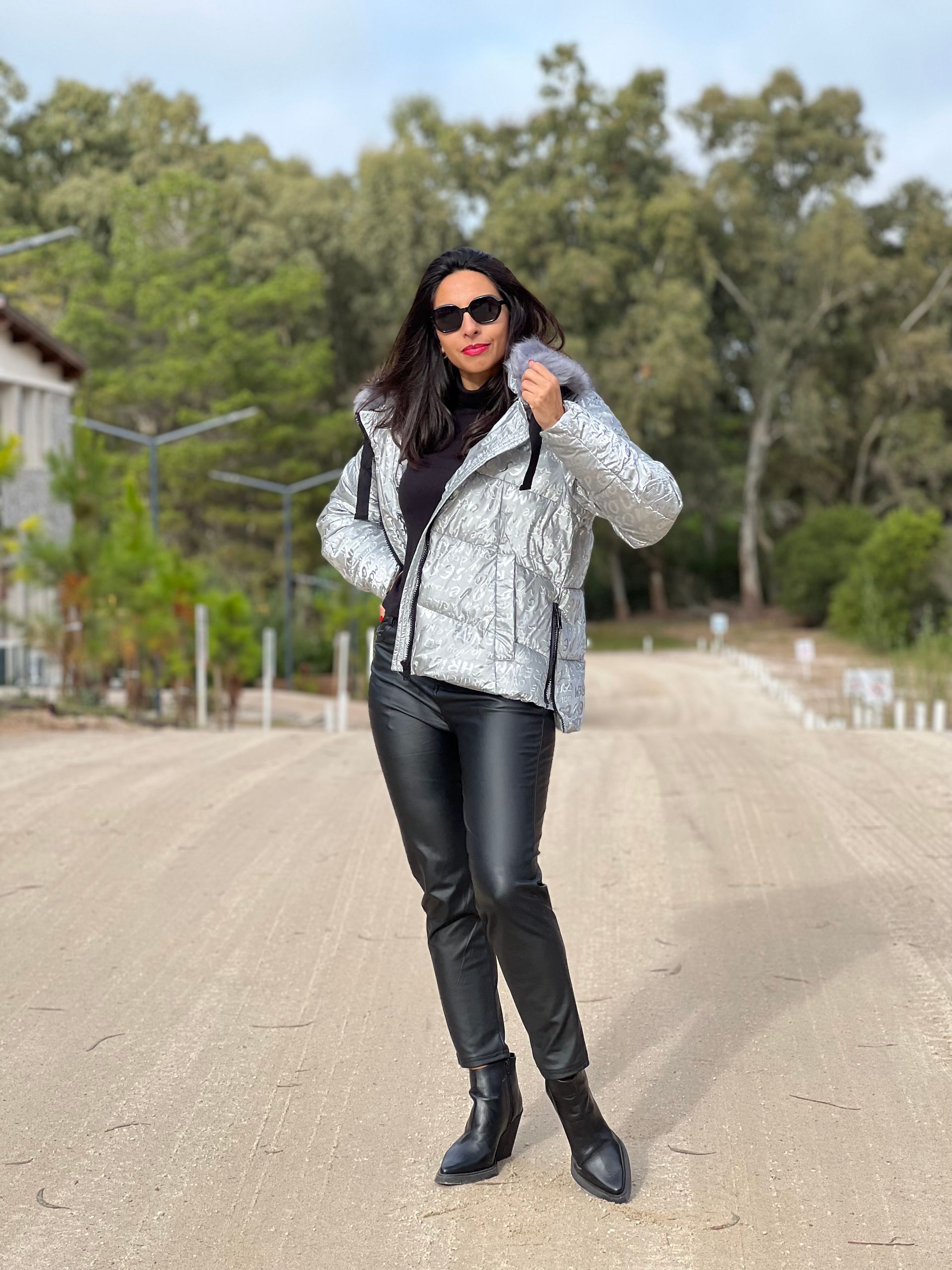 Mujer con gafas de sol ajustándose una chaqueta plateada con lentejuelas, un top negro y pantalones de cuero al aire libre.